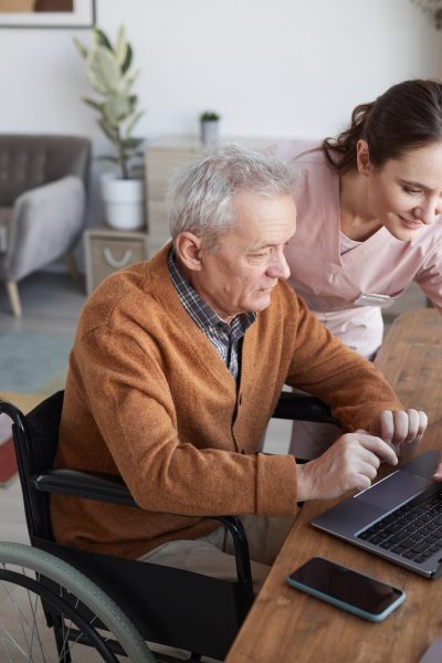 Portrait of senior man in wheelchair using laptop at retirement home with nurse assisting him, copy space